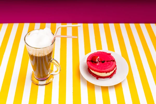Beyaz ve sarı çizgili bir masada bir fincan kahvenin yanında ahududu turtası..