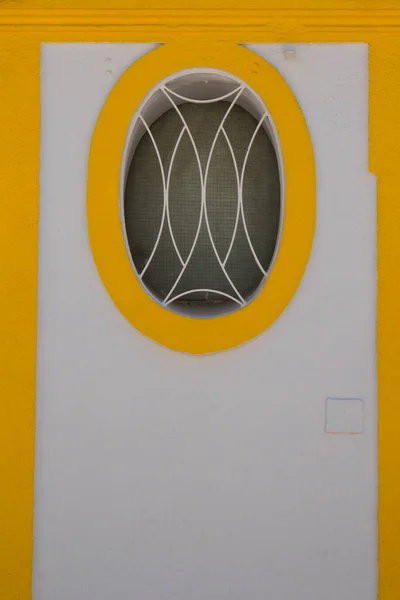 White wall of a house with oval window with a grid. Window lined by yellow painted frame. Next yellow lines. Vila Real de Santo Antonio, Algarve, Portugal.