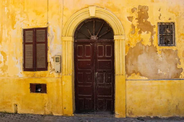 Yellow old facade of a house with wooden brown gate with an arch. Window with a shutter. Street in former portuguese fortress in El Jadida, Morocco.