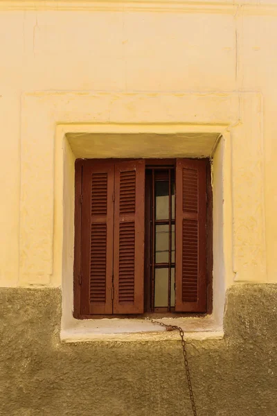 Yellow wall of a house with a window with partly opened brown shutter. Rusted chain hanging from the window. El Jadida, Morocco.