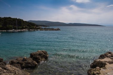 Kayalar Adriyatik Denizi kıyısında. Sakin su küçük dalgalar ile. Orman diğer yan ve arka planda dağlar. Bulutlu gökyüzü. Şehir Krk, Island Krk, Hırvatistan.