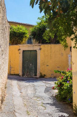 Dar yıpranmış yol ve kaldırım, bitkilerle kaplı. Sarı cephe eski kapısı olan bir ev bir çit. Mavi gökyüzü. Geleneksel köy dağlarda Archanes, Crete, Yunanistan.