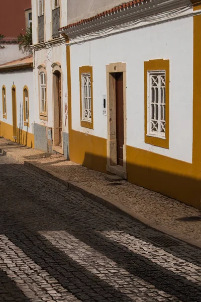 Road made of cobblestones with a crosswalk, lined by houses with white facade and yellow details. Silves, Portugal.