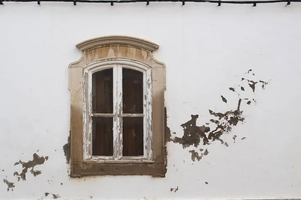Peeled of wooden frame of the window with a small arch, which is in a stone ornate frame. White facade of the house with weathered peeled off parts, creating a kind of decor. Tavira, Portugal.