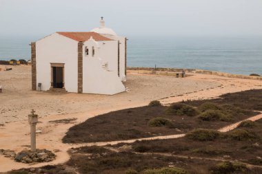 Kumlu toprak ve Atlantik Okyanusu ile manzara. Beyaz kilise çan ve bir haç ile. Bulutlu gökyüzü. Kale Sagres, Portekiz.