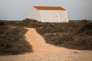 Kale uçurumlarda Çalılar sonbaharda kuru. Beyaz turuncu bir çatı ile bina. Bulutlu yağmurlu gökyüzü. Sandy patika. Sagres, Portekiz.