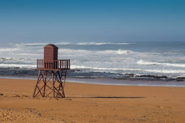 Golden clean sand of the beach. Wooden lifeguard tower. High tide, waves with white foam. Empty beach in the autumn. Horizon of the Atlantic ocean. Blue sky. Beddouza Beach, Morocco.