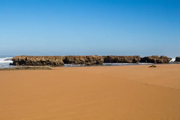 Golden sandy beach with little rocks on the coast. Wild water of the Atlantic ocean with waves. Clear blue sky. Horizon in the background. Beddouza beach, Morocco.