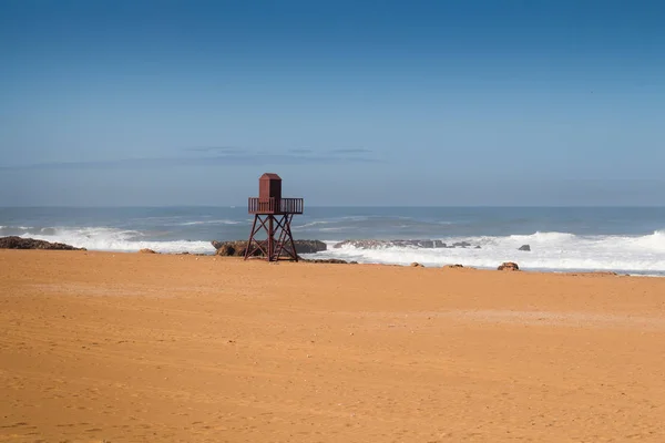 Golden clean sand of the beach. Wooden lifeguard tower. High tide, waves with white foam. Empty beach in the autumn. Horizon of the Atlantic ocean. Blue sky.