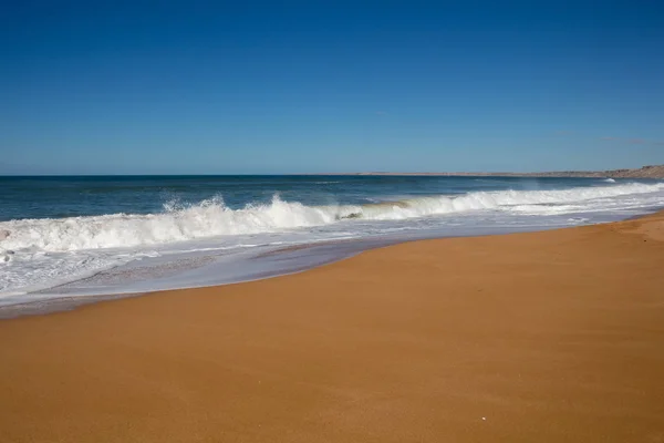 Clean dark yellow sand of the autumn beach. Deep blue color of the Atlantic ocean water with foamy waves. Horizon in the background. Clear blue sky. Lalla Fatna beach (Safi). Morocco.