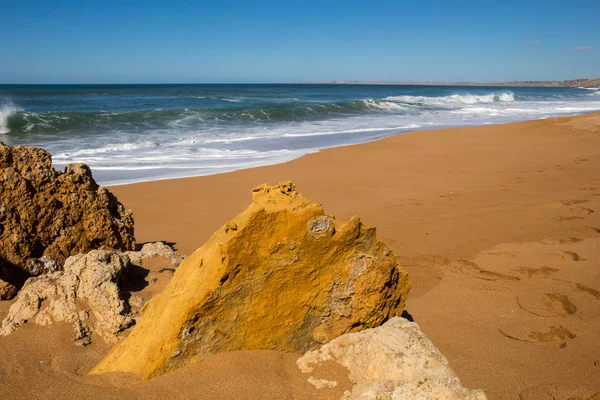 Rocks on the clean beach with dark yellow sand. Foamy waves of the Atlantic ocean. Horizon in the background. Clear blue sky. Lalla Fatna beach (Safi), Morocco.