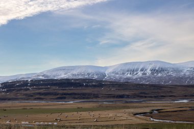 Büyük tarlalar ve otlaklar. Arkasında kar olan dağlar. Vadideki sığ nehir. Beyaz bulutlu mavi gökyüzü. Siduvegur Bölgesi, Kuzey-Batı İzlanda.