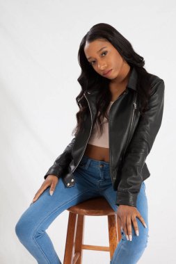 Deri ceketli, kot pantolonlu, tahta bir taburede oturan güzel siyah kadın.