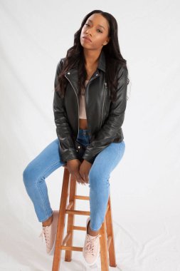 Deri ceketli, kot pantolonlu, tahta bir taburede oturan güzel siyah kadın.