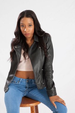 Deri ceketli, kot pantolonlu, tahta bir taburede oturan güzel siyah kadın.