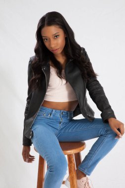 Deri ceketli, kot pantolonlu, tahta bir taburede oturan güzel siyah kadın.