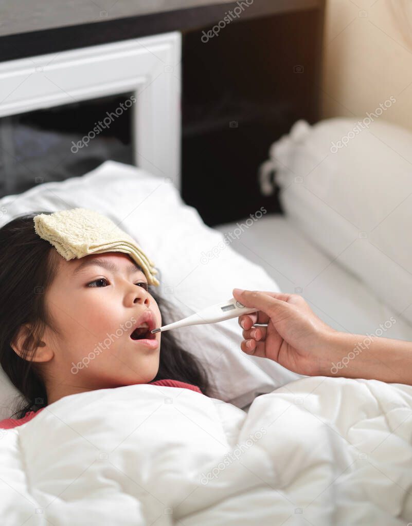 Asian mother measuring temperature girl with digital thermometer in her mouth on bed at morning time, Sick child have cool towel for reduce high fever,  Selective focus, Healthy and infection concept