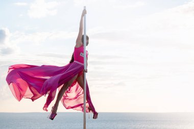 Kumsaldaki uzun mavi kumaşlı sıska kadın. Yazın gökyüzü arka planı. Doğada Polonyalı dansçı. Denize ve gökyüzüne karşı kadın direk dansçısı.