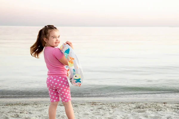 Toddler girl in pink clothes stands on seashore and holds inflatable toy. Little girl lookihg at the camera on sunset beach. Childrens games on the beach summer day.