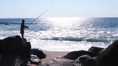 Okyanus kıyısında bir balıkçı. Kamera. Bir balıkçı okyanusa bir olta atıyor. Arkadan manzara. Güneşli deniz manzarasının arka planında bir balıkçının silueti.