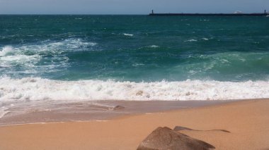 Kum kıyısı ve kıyıdan yükselen resimli dalgalar. Derin, yumuşak bir su renginde Atlantik Okyanusu. Arka planda bir fener var..