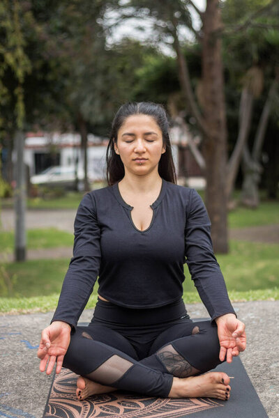 Женщина в черной одежде медитирует в парке на фоне деревьев
