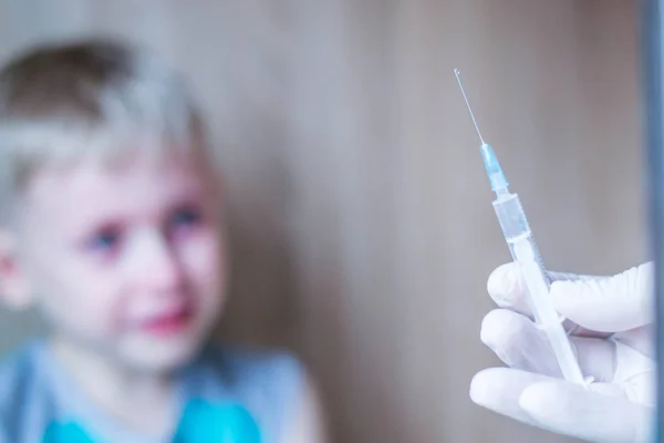 Çocuk mavi gözlü sarışın. Korku ve umutsuzluk hissediyor. Enjeksiyonun tıbbi prosedüründen çok korkuyorum. İğne ler için şırıngaya baktığında ağlıyor. Turkuaz tişört giymiş bir çocuk. Beyaz eldiven el bir çözüm şırınga tutar