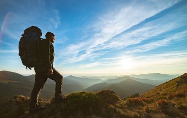 Kayanın üzerinde kamp çantasıyla duran adam. Gün batımının resmedildiği.