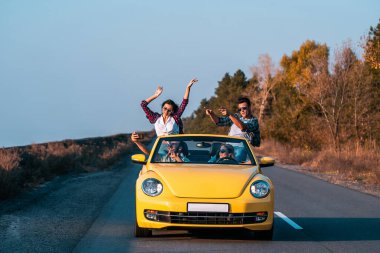 Genç arkadaşlar üstü açık sarı bir arabayla seyahat ediyorlar.