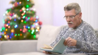 Gözlüklü yorgun emekli Noel ağacının yanında bir kitap okudu.