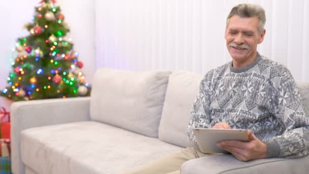老人坐在沙发上 用一块靠近圣诞树的石碑与人交谈 — 图库视频影像