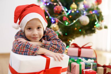 Gülümseyen çocuk Noel ağacının yanında bir hediye kutusuyla oturuyor. 