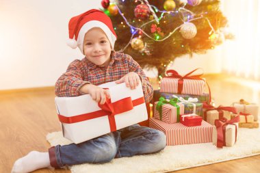 Mutlu çocuk güneşli bir arka planda Noel ağacının yanında bir hediye kutusuyla oturuyor.