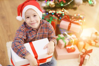 Mutlu çocuk güneşli bir arka planda Noel ağacının yanında bir hediye kutusuyla oturuyor.
