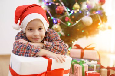 Gülümseyen çocuk, elinde bir hediye kutusuyla güneşli bir arka planda Noel ağacının yanında oturuyor.