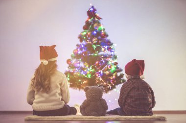 Yumuşak oyuncaklı bir kız ve oğlan Noel ağacının yanında oturuyorlar.