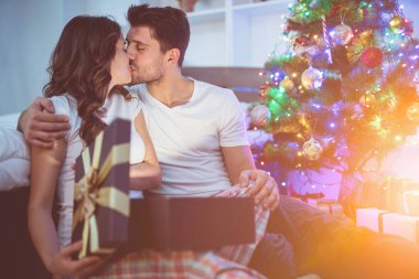 Öpüşen çift Noel ağacının yanında bir hediye kutusuyla oturuyor.