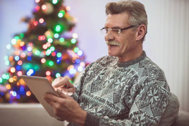 Tabletli yaşlı adam Noel ağacının yanında oturuyor.