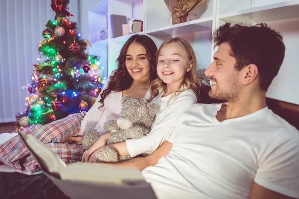The happy family lay on the bed and read a story near the christmas tree