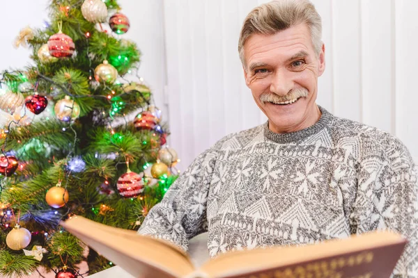 The old man hold a book near the christmas tree