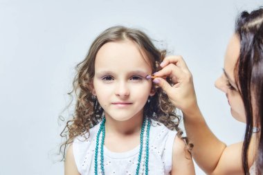 Anne küçük kızına makyaj yapıyor. Stüdyoda beyaz sırtlı bir fotoğraf çekimi.