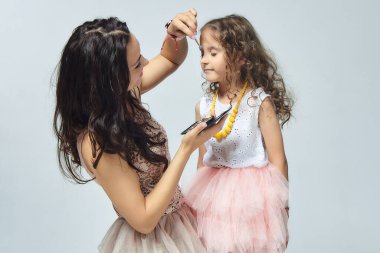 Anne küçük kızına makyaj yapıyor. Stüdyoda beyaz sırtlı bir fotoğraf çekimi.