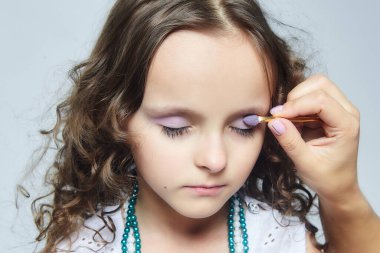 Anne küçük kızına makyaj yapıyor. Stüdyoda beyaz sırtlı bir fotoğraf çekimi.