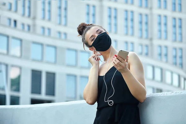 beautiful girl in a mask, posing in the city, holding a smartphone and listening to music with headphones.