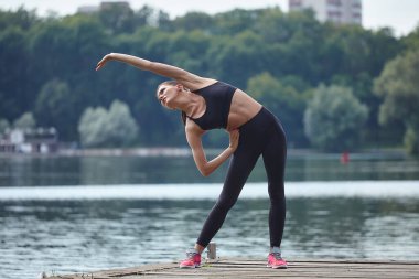 Spor giyim sektöründe çekici bir kız şehir parkındaki ahşap iskelede jimnastik yapıyor..