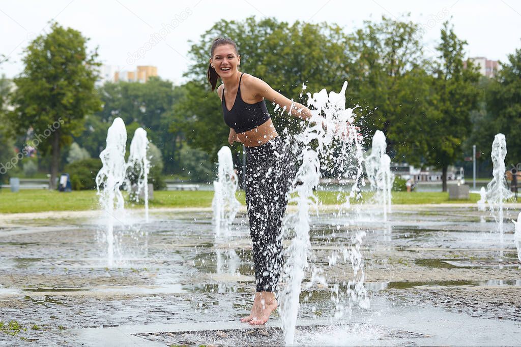 Attractive girl happily plays with fountain water stabs in city park.