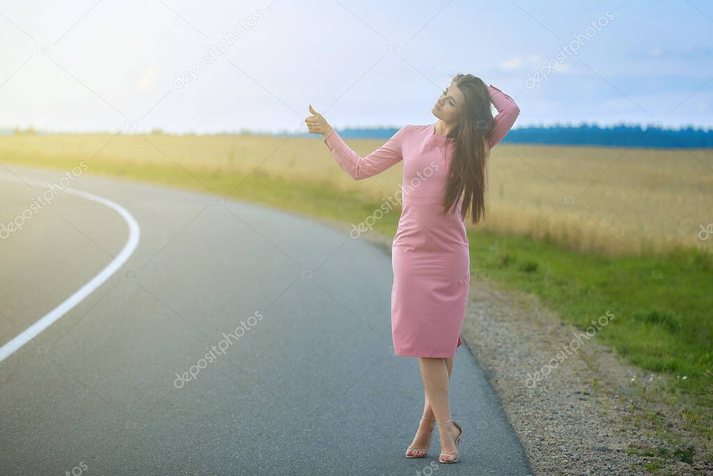 beautiful girl in a pink dress stands on the road with a raised hand in anticipation of a passing car