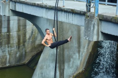 Sporcu bir insan jimnastik ipinde tehlikeli egzersizler yapar. Arka planda su akıntısı olan bir baraj var.