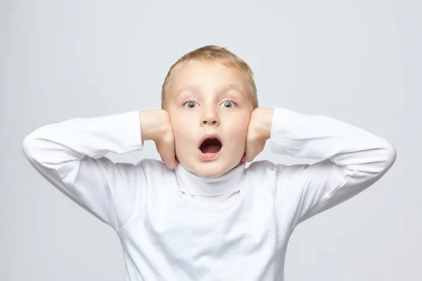 Young blond boy covers his ears with his hands and opens his mouth in surprise