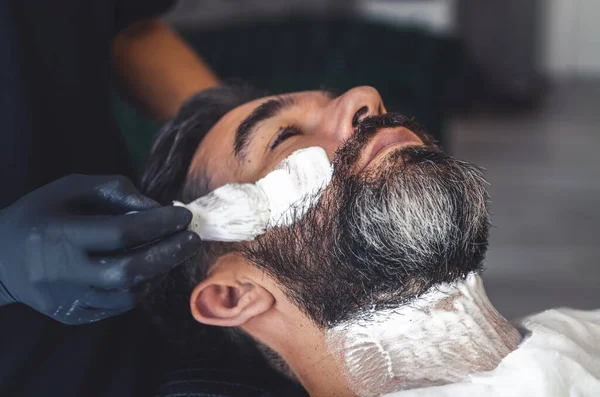 barber with latex gloves applying cream with a brush to a client to shave the beard